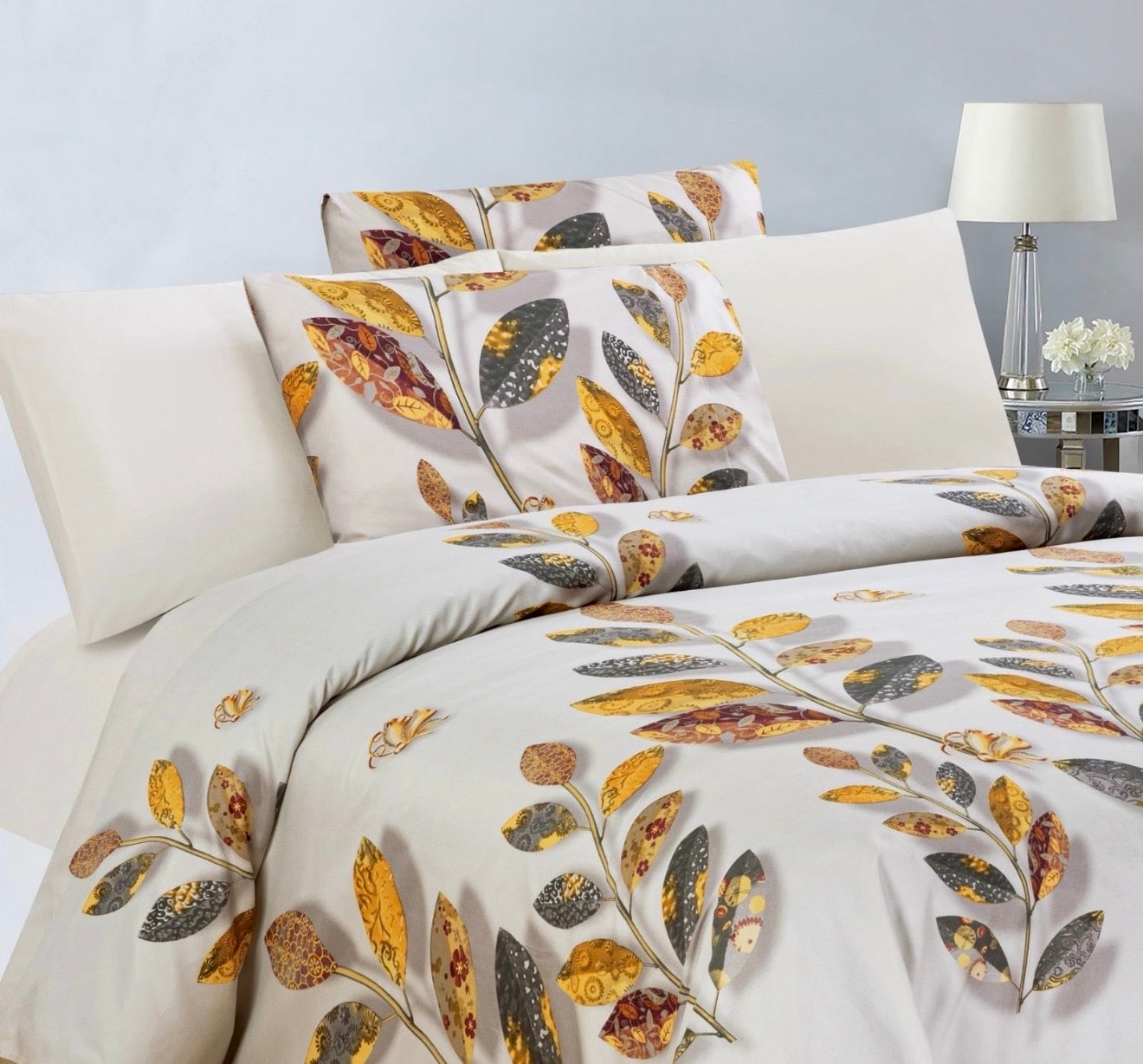 A bedding set on a bed featuring a cream-colored Bedsheet and matching pillowcases with a detailed autumn leaf pattern in yellow, brown, and grey. The bed is styled in a bedroom setting with a bedside lamp and flowers visible on the right.